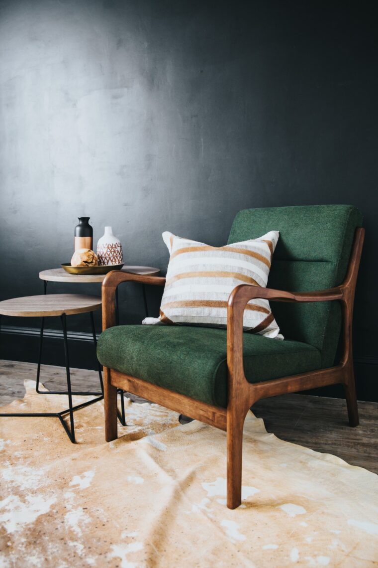 A wooden chair with dark green cushions and a striped pillow on it. There are two side tables next to the chair and a spotted rug below.